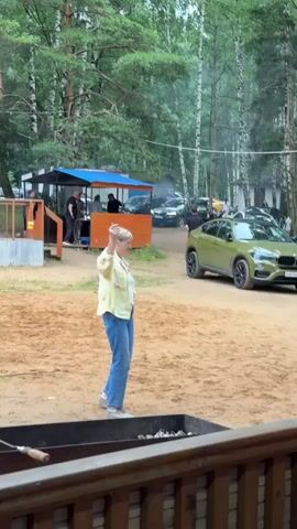 A woman with short blonde hair, wearing a light-colored jacket over a white t-shirt and blue jeans, stands in a sandy outdoor area. She is facing left and appears to be waving or gesturing with her right arm. In the background, a blue and orange shelter is visible, with several people standing or sitting under it. Cars are parked behind the shelter. A green SUV is positioned to the right of the woman. In the foreground, the edge of a dark metal barbecue grill is visible. The environment appears to be a wooded area.
