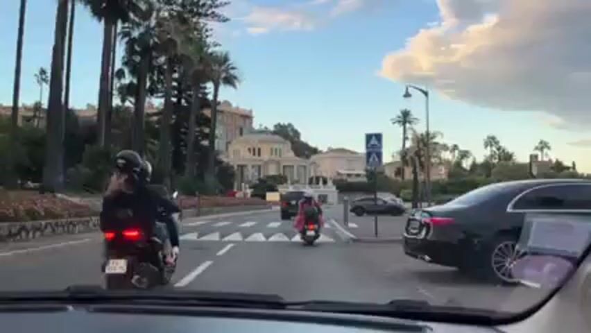 The view from inside a car shows two people on motor scooters driving ahead across a crosswalk.  They are wearing helmets. The background shows palm trees, a building with classical architecture, and other cars parked along the street. The sky is blue with wisps of white clouds. There is music playing in the car.