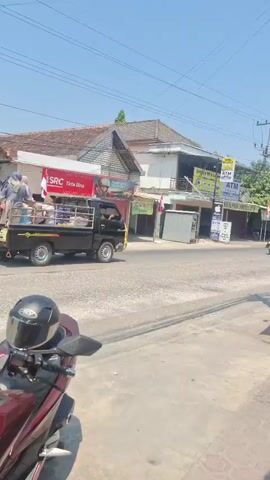 A daytime, eye-level, medium shot captures a street scene with a busy road. In the foreground, the front of a red motorcycle is visible, with its handlebars and helmet in focus. The middle ground shows a paved road with several vehicles. A black pickup truck is driving away from the camera, loaded with goods and people. Two motorcycles, each carrying two people, are also traveling in the same direction. To the left of the road, a building with a red and white sign reads "SRC". To the right, a utility pole is covered in numerous wires and a blue tarp is erected on the sidewalk. The background features more buildings and a clear blue sky.