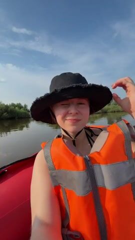 This video shows a group of people in a red inflatable boat on a river. The first shot is a selfie of a young woman wearing a black bucket hat and an orange life vest, smiling at the camera. She is in the front of the boat. The second shot shows the boat moving on the water with three other people in it. A man in a green cap and a blue towel is in the center of the boat, facing away from the camera. A man wearing a life vest is on the left, and a woman wearing an orange life vest is on the right. The background shows a tree-lined shore and a blue sky with clouds. The water is calm and reflects the sky. The boat is leaving a wake behind it. The overall atmosphere appears to be one of a leisurely boat trip on a sunny day.