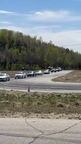 The video shows a parade of approximately nine cars driving along an asphalt road with a turn. The cars are various colors including silver, light blue, and white. Many of the cars have white flags attached to them. The cars are driving from right to left on the screen. The surrounding area includes green trees, a small hill, and dry grassy areas. There is also a red and white traffic cone along the side of the road. The sky is blue with some white clouds. The road has cracks and worn areas. There are some other cars parked in the background.