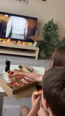 A young person sits with their back to the camera, watching a television screen that displays a scene from a show or movie. In front of them, on a white coffee table, is a box of pizza with several slices remaining, a can of Pepsi, and a small container of dipping sauce. The young person is holding a slice of pizza and appears to be eating it. The room is dimly lit, with string lights on the television stand creating a warm ambiance. A large plant in a white pot is visible to the right of the television. The floor is light-colored wood.