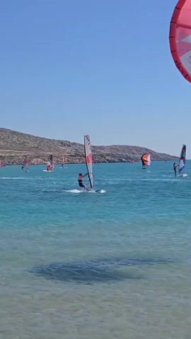 The video shows a bright, sunny day with a clear blue sky and calm turquoise waters. The scene is set at a beach with a low, rocky coastline in the background. The primary activity depicted is windsurfing, with several individuals actively engaged in the sport. One windsurfer in the foreground is wearing a red and black outfit and is captured in mid-action, their sail angled to catch the wind. To their left, another windsurfer is visible further out. In the middle ground, multiple other windsurfers are spread across the water, their sails varying in color, including orange and red. A large, pink and white sail, possibly from a kite, is prominently featured in the upper right portion of the frame, partially obscuring the sky. The water exhibits a sandy bottom near the shore, creating a shallow area with visible shadows, which transitions to deeper blue further out. The overall atmosphere is one of recreational activity on a beautiful day by the sea.