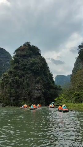 The video shows several small boats carrying people navigating a river surrounded by mountains. The people are wearing orange life vests. The boats are moving through the water, creating small ripples. The mountains are covered in lush green vegetation and are a combination of rocky cliffs and gentle slopes. The sky is overcast with heavy clouds. The scene conveys a sense of exploration and natural beauty.