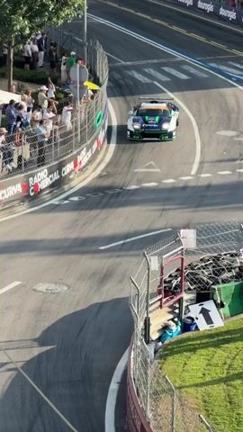 A race car is shown from a high angle as it navigates a turn on a track. Spectators are visible behind a fence on the left side of the frame. The car is primarily white, blue, and green, with various sponsor logos. The track is paved, with white lines indicating the racing surface. There are also barriers and safety fences along the track. The sun is casting shadows, suggesting it is daytime and the sun is low in the sky.