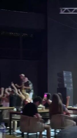 The video shows a dimly lit indoor venue with a stage at one end. On the stage, two musicians are performing. One man, wearing a hat and a patterned shirt, is playing a guitar. Another man, wearing a floral shirt and white pants, is playing a saxophone. They are both standing and facing the audience. Several audience members are visible in the foreground, seated at tables. Some are clapping or raising their hands in the air, suggesting they are enjoying the performance. The environment appears to be a bar or restaurant with a stage set up for live music. The lighting is moody, with spotlights on the performers.