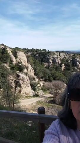 A woman with gray hair and sunglasses stands facing the camera in an outdoor setting. She is wearing a light pink sweater. Behind her is a scenic landscape with rock formations and scattered trees. A road winds through the terrain. In the distance, there is a view of fields and possibly a town under a bright sky. A wooden fence is in the foreground near the woman. As the camera angle slightly shifts, a castle tower is revealed in the background.