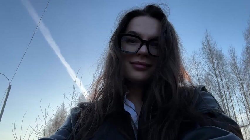 A woman is taking a selfie outdoors on a sunny day. She has long brown hair, is wearing glasses and a black leather jacket over a white shirt. She is smiling at the camera and runs her hand through her hair. Behind her is a row of bare trees and the sky is visible with a contrail. There is also a road with traffic signals and cars in the background.