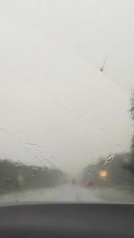 A windshield wiper sweeps across a car windshield during a rainstorm. Water streams down the glass, obscuring the view of the road ahead, which is lined with trees and other vehicles. The sky is overcast and white, creating a hazy atmosphere. The scene appears to be a highway or a similar type of road.