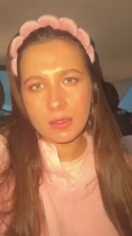 The video shows a young woman with long, dark brown hair wearing a pink hooded sweatshirt and a pink fuzzy headband with floral accents. She is sitting in the back seat of a car, facing the camera. The woman's facial expressions change from closed eyes to looking around. There is a child and bags visible in the back of the car. The setting appears to be overcast.