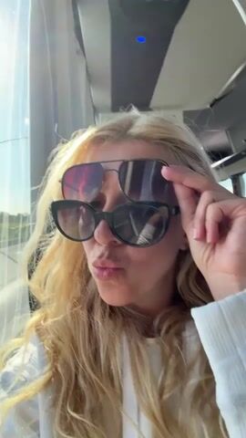 A blonde woman with long curly hair is sitting inside a vehicle. She is wearing two pairs of sunglasses, one over the other. She is looking to her left and pouting her lips. Her hand is raised, and she is adjusting the top pair of sunglasses. She then looks forward and smiles at the camera. The background shows a window with a blurry view of the outside and the interior of the vehicle's ceiling.