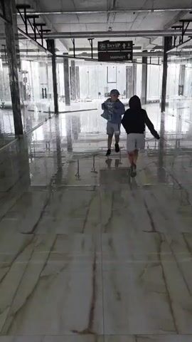 Two children are walking across a wet, reflective floor in what appears to be an unfinished or empty indoor space. The floor is tiled with a marble-like pattern. Above them, suspended from the ceiling, are signs indicating "SKY UP" and a market. The space has a concrete ceiling with exposed pipes and metal beams. Large windows line the background, showing an overcast or rainy outdoor environment. The children are dressed casually, one in a light-colored jacket and the other in a dark hooded jacket. They are walking away from the camera, towards the signs.