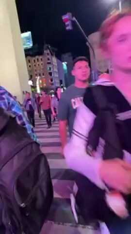 The video shows a nighttime street scene with people crossing a crosswalk. The crosswalk is illuminated with a pink light. There's a tall obelisk-like structure on the left side of the frame. In the background, there are buildings and streetlights, and what appears to be a Ferris wheel. People are walking in various directions across the crosswalk. Some are talking.