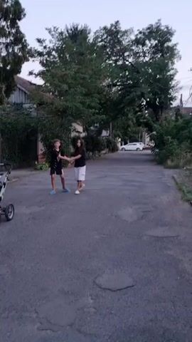 An adult woman wearing a white t-shirt and black pants with a pink backpack walks away from the camera on an asphalt road. Ahead of her, two children, a boy and a girl, are holding hands and standing next to a stroller. The boy wears a black t-shirt and dark shorts, and the girl wears a black t-shirt and white shorts. The girl is pushing the stroller, which contains a baby. Trees and buildings line the road, and a white car is visible in the distance. The scene appears to be outdoors during daylight.
