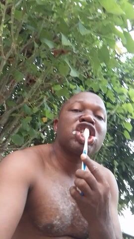 A man with dark skin is seen brushing his teeth in front of green foliage. He is shirtless and faces the camera while holding a blue and white toothbrush. The camera angle is low, looking upwards at the man, and the background consists of dense green leaves and branches. He appears to be focused on his oral hygiene.