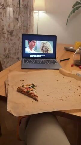 A close-up shot shows a laptop on a wooden table. The laptop screen displays a video with two people talking, and Russian subtitles. To the left of the laptop, a slice of pizza sits on brown paper. To the right, a wine glass and a utensil are visible. In the background, a lamp and a curtain are partially visible. The video on the laptop screen then changes to show Donald Trump speaking at a podium in front of a crowd, with "BATTLE CREEK, MICHIGAN" and "C-SPAN" displayed on the screen. The Russian subtitles continue.