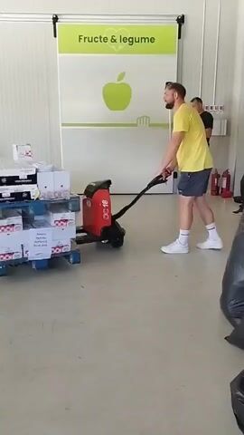 A man in a yellow t-shirt and blue shorts operates an electric pallet jack loaded with boxes. He is pulling the pallet jack towards the left side of the frame. The pallet jack has a red body and a black handle. The boxes on the pallet are stacked high and wrapped in clear plastic. In the background, another man in a dark t-shirt and shorts stands near a large green sign that says "Fructe & legume" with a picture of an apple. There are shelves and other industrial equipment visible in the background. The floor is concrete.
