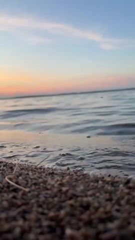The video is a close-up, low-angle shot of a sandy shoreline at sunset. Gentle waves lap at the shore, reflecting the warm hues of the sky, which transitions from pale yellow near the horizon to a soft blue overhead, with streaks of pink and orange clouds. The sand is composed of small pebbles and grains. The sun is visible on the horizon, a bright orange disc. A thin, dry blade of grass lies on the sand in the foreground, slightly out of focus. The overall mood is peaceful and serene.