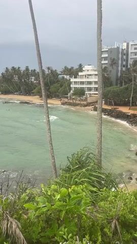 The video shows a view of a tropical beach and ocean from a high vantage point, partially obscured by palm trees and lush green foliage in the foreground. The ocean water is a vibrant turquoise color, with small waves breaking near the shore. A sandy beach stretches along the coastline, lined with palm trees and a few buildings, including a multi-story white hotel. There are two small boats visible in the water, one with people in it. The sky is overcast, suggesting a cloudy day. The overall scene is serene and picturesque, evoking a sense of a tropical getaway.