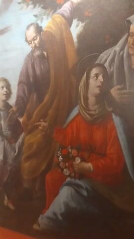 This is a photograph of a painting, possibly a religious depiction, as suggested by the solemn expressions and the presence of figures in robes. The painting is oriented vertically and appears to be of a significant size, given the detail visible.

The subjects are several individuals, rendered in a classical artistic style. In the foreground, two figures appear to be looking towards the viewer, with one possibly a younger person with curly hair. Behind them, a person in a purple garment is partially obscured by a large, flowing yellow and brown cloak. Above this, a central figure, likely female, is dressed in a vibrant red robe and a head covering, holding a bouquet of flowers. Her gaze is directed upwards. At the very top of the frame, another figure is visible, appearing to be an older person, also with a muted colored robe and a relaxed, almost sleeping posture.

The scene seems to be set outdoors, suggested by the presence of some foliage with red berries on the left side of the painting. The lighting is soft and diffused, hinting at a natural light source, possibly daytime, but without strong shadows to indicate a specific time. The overall mood appears to be one of quiet contemplation or solemnity. There is no visible text in the image. The context provided indicates the location as Peccioli, Italy, suggesting this artwork may be displayed in a museum, church, or public space within the town.