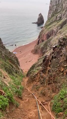 The video shows a person climbing a steep, rocky slope towards a beach. The slope is flanked by rocky cliffs covered with vegetation. Two ropes, one blue and one orange, are stretched along the slope, apparently for assistance in climbing. The person climbing is wearing a red jacket and a backpack. The beach below is sandy and there is a body of water beyond it. A prominent rock formation rises from the water. The sky is overcast.