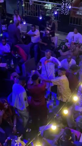 The video depicts a lively gathering in a nightclub setting. Several men are seen standing around tables, some drinking and interacting with each other. One man in a white traditional outfit appears to be filming with his smartphone, while another man in a maroon outfit is adjusting his head covering. The scene is illuminated with blue lighting, and tables are filled with bottles of drinks. Overall, the video captures a social atmosphere with individuals enjoying themselves in a nightclub environment.