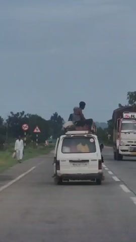 A white van is traveling on a paved road. The van is overloaded with luggage on its roof. A person is sitting on top of the luggage on the roof of the van. Another person is visible inside the van. The road is flanked by trees and greenery. There are road signs on the side of the road. Another vehicle, a truck, is visible further ahead on the road. The sky is overcast.