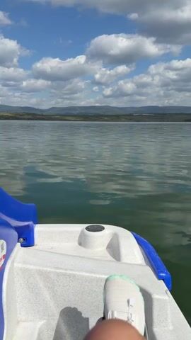 A first-person perspective video shows the front of a pedal boat on a calm lake. The boat is white with blue accents. The water is green and reflects the sky and clouds. In the distance, a tree-lined shore can be seen. The sky is blue with fluffy white clouds. A person's leg, wearing a white sneaker, is visible in the bottom right corner of the frame. The boat appears to be moving slowly through the water.