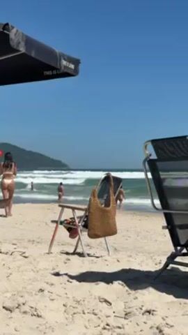 The video shows a sunny beach scene.  In the foreground, a white folding chair with a brown woven bag hanging on it sits in the sand. To the right of the folding chair is a black and silver beach lounge chair. A large black beach umbrella with white lettering saying "THIS IS LIVING" is partially visible at the top left corner of the frame. In the background, people are walking and standing in the ocean water, which is choppy with waves. A green, hilly landmass is visible in the distance. The sky is a clear, bright blue.