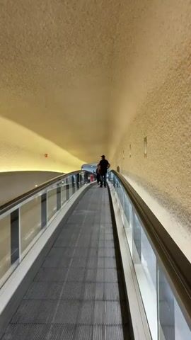 The video shows a first-person perspective traveling on a moving walkway. The walkway has a textured gray surface with ridged patterns. To the left and right, there are transparent glass barriers with silver railings. The ceiling is a beige, textured material that curves downwards, creating a tunnel-like effect. Circular light fixtures are embedded in the wall on the right. In the distance, several people are visible walking on the walkway. The lighting is warm and appears to be coming from the ceiling and possibly the sides. The overall environment suggests an indoor public space like an airport terminal or a large shopping mall. The perspective is forward-looking, indicating movement along the walkway.