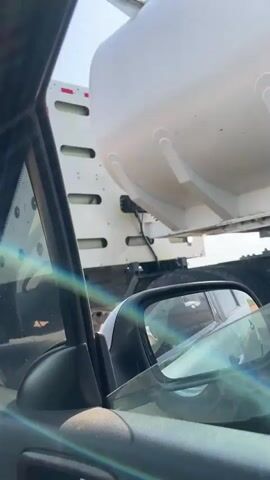 The video shows a person in a car, recording through the car window. The camera angle focuses mostly on the side window of the car. At the beginning of the video, a white truck with a white object containing black circles is seen. The camera then focuses on the sky.  The camera then shows a blue truck carrying cargo.  The rest of the video shows the blue truck. The scene appears to be on a highway.