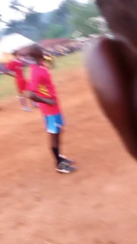 People are on a dirt field. Some players are in yellow and blue uniforms, while others are in red uniforms. A person in red moves toward the left side of the frame. The video is shaky. In the background, there is a tent and some buildings. There are other people on the field and along the edge. The video ends abruptly.