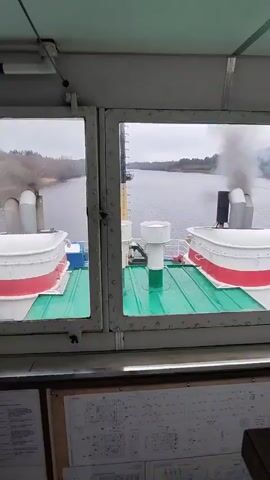 The video is shot from inside what appears to be the bridge of a boat or ship. The view through the windows shows a wide river or body of water. The landscape is overcast, with trees lining the banks on either side. The camera is stationary. The main focus is on the rear of the ship itself, where two sets of smokestacks are visible. One set is emitting visible smoke. The stacks are painted white with red trim and are situated on a green surface. Between them, a mast rises with a Russian flag. Below the windows, there is a schematic diagram or technical drawing attached to the wall. The overall impression is a still, quiet scene captured from a vessel moving along a river on a cloudy day.