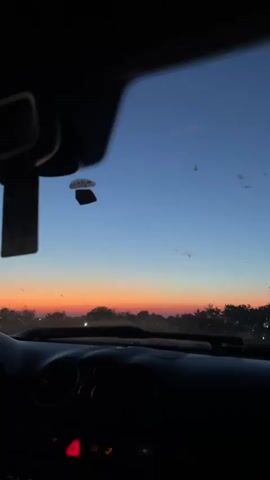 The video is shot from inside a car, looking out through the windshield at a sunset. The sky is a gradient of blue to orange and pink. Silhouettes of trees and some buildings are visible on the horizon. The dashboard of the car is visible in the foreground, with some buttons and a rearview mirror. There are some specks on the windshield, possibly dirt or water spots.