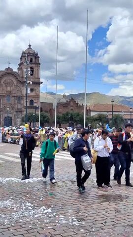 A group of musicians is parading through a plaza. They are playing drums and flutes. The musicians are mostly men, but there is also a young boy playing a snare drum.  There is a boy wearing a green sweater holding confetti poppers.  They are marching on a paved square strewn with confetti. The buildings in the background are old and made of stone. There is a church with a tall tower in the background. There are other people in the background, possibly watching the parade. The sky is cloudy, but there is some blue sky visible. There are mountains in the distance.