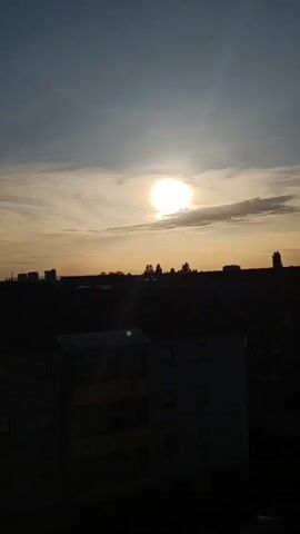 The video shows a sunset over a cityscape. The sun is a bright orb in the sky, partially obscured by clouds. The sky is a gradient of orange, yellow, and blue. Silhouettes of buildings and trees are visible against the horizon. The video is shot from a high vantage point, likely a window or balcony, looking down on the scene. There are no people visible in the video. The overall mood is calm and serene.