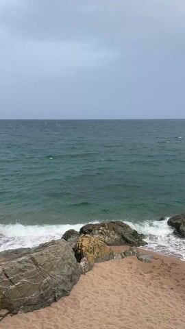 The video shows a view of the ocean from a rocky beach on an overcast day. The sea is a deep teal color with gentle waves breaking against the shore and rocks. The beach is sandy with scattered rocks of varying sizes, some covered in moss or seaweed. The sky is uniformly gray. The overall mood is calm and serene.