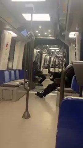 The video shows the interior of a mostly empty subway car. Several passengers, all appearing to be male, are seated along the sides of the car. The seats are upholstered in blue, and the interior is clean and well-lit with white and blue tones. There are vertical metal poles for standing passengers to hold on to. The subway car appears to be in motion, as a slight motion is visible in the video. The scene suggests a quiet, perhaps late-night or early-morning, commute.