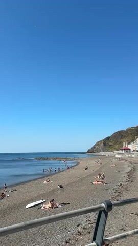 The video shows a pebble beach with a clear blue sky and calm ocean. Several people are scattered on the beach, some sunbathing on towels and others in the water. A white surfboard lies on the sand. A metal railing runs across the foreground. The scene is peaceful and appears to be a sunny day at the seaside.