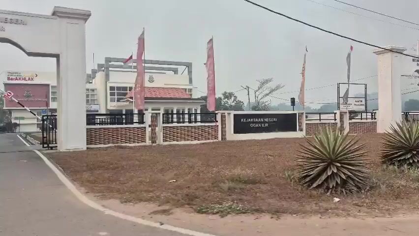 The video shows a passing view of the Kejaksaan Negeri Ogan Ilir, which appears to be a courthouse or prosecutor's office in Indonesia. The building is visible on the left, with flags flying in front. A large archway with a gate serves as the entrance. The foreground features dry, brown grass and several spiky plants. The overall environment appears to be somewhat dry and dusty, with a cloudy or hazy sky. There are no people clearly visible in the video. The camera is moving, suggesting it is being filmed from a vehicle.