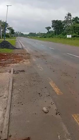 The video shows a white Toyota pickup truck driving away on a wet, two-lane road in what appears to be a rural or semi-rural area. The road has a combination of solid white lane markers and dashed yellow center lines. The shoulders of the road are grassy, and there are trees visible in the background along with a cloudy sky. There is a pile of gravel or asphalt on the left side of the road near the beginning of the video. The road surface itself appears to be patched and has some debris scattered on it.