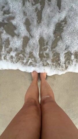 A person stands in shallow water at the beach. The camera looks down at their legs and feet. Waves with white foam wash around the person's feet. The person has a dark tan and blue toenail polish. Some sand sticks to their legs.
