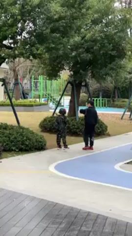 In a park-like setting with trees, a small playground, and landscaped areas, two young children are walking along a blue and white path. One child is wearing a camouflage outfit and helmet, while the other is dressed in black athletic wear with red shoes. The child in camouflage is walking and gesturing with their arm raised. The child in black is walking and looking toward the playground in the background. A person can be seen swinging on a swing set in the playground. The weather appears overcast. The scene is filmed at a medium distance, providing a clear view of the children and their immediate surroundings.