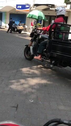 A three-wheeled vehicle with a black cargo bed is moving forward on a paved street. The vehicle is branded "VIAR" and has a license plate that reads "H 6122 WSP." A person wearing a red shirt and a white helmet is driving the vehicle. Another person, partially visible, is seated in the cargo bed. The cargo bed contains bags and other items. In the background, buildings and people can be seen, suggesting an urban or market environment. The lighting indicates it is daytime.