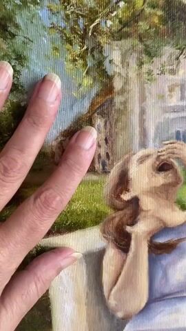 A hand with light skin and short, unpainted fingernails is placed over a painting. The painting depicts a woman in a light blue dress sitting on a stone bench outdoors, laughing. She is leaning back, with her head tilted upwards and one hand covering her mouth and eyes. The background of the painting shows trees, grass, and buildings. After the hand moves, the painting is shown in full, framed, on a white surface with a glass of paintbrushes and a book.