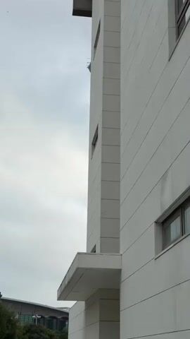 The video shows a tall, modern building. A cable or rope hangs down from near the top of the building, with the top portion appearing to be a cylindrical observation deck. The building's exterior is a light gray color, and it tapers slightly toward the bottom. The background sky is overcast.  Other smaller structures are visible at the base of the tower. The camera pans upward along the length of the building.