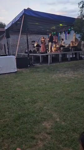 A live band performs on an outdoor stage. The stage is covered by a blue tarp and illuminated by green and purple stage lights. A male vocalist stands center stage, playing an acoustic guitar. To his left, a female vocalist sings into a microphone. Behind them, other band members play instruments, including drums and a keyboard. A backdrop depicting a city skyline is visible behind the performers. The stage is set up on a grassy lawn, with trees and a stone building visible in the background. The sky is overcast, suggesting it is either early morning or late evening. A white covered object, possibly a speaker, is positioned to the left of the stage.