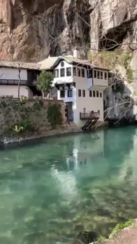 The video shows a complex of buildings located next to a turquoise river and underneath a steep cliff. The camera pans from right to left, revealing a white, multi-story structure with black trim and a balcony, followed by a stone wall, and then another building constructed from brown stone. The river appears clear and shallow, with some visible rocks on the riverbed. There is light vegetation, including small trees and grass, dotting the landscape around the buildings. The overall scene suggests a historic or culturally significant site.