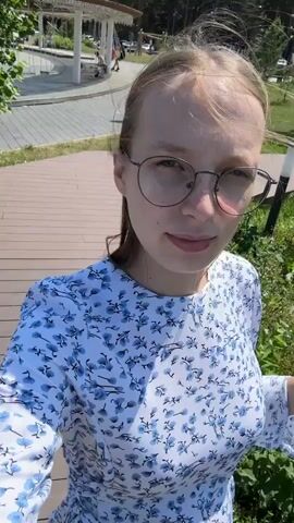 A young woman with blonde hair and glasses is taking a selfie outdoors. She is wearing a white dress with a blue floral pattern. She is standing on a wooden walkway next to some greenery. In the background, there is a park with trees, a fountain, and people walking. The sun is shining.