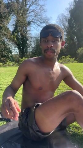 A young, dark-skinned man wearing dark sunglasses and black leather shorts sits cross-legged on a black blanket in a grassy park on a sunny day. He has a mustache and short, dark hair. The man adjusts his sunglasses. There are trees visible in the background.
